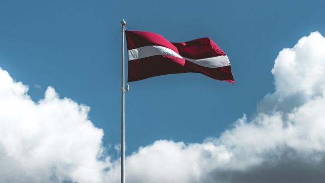 Latvijas karogs mastā, fonā zilas debesis ar mākoņiem