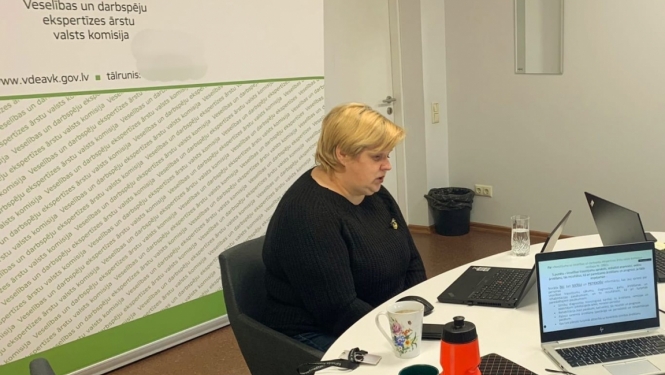 galds, pie galda sēž sieviete, uz galda dators, ūdens pudele, piezīmju grāmata