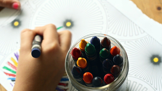 Bērns krāso zīmējumu ar krītiņiem. Fonā trauks ar dažādas krāsas krītiņiem