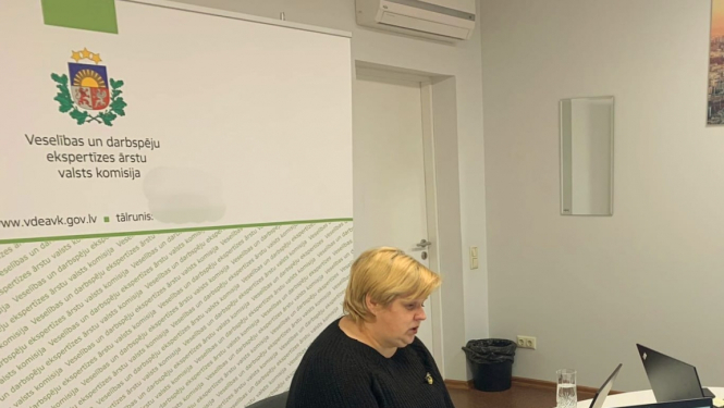 galds, pie galda sēž sieviete, uz galda dators, ūdens pudele, piezīmju grāmata