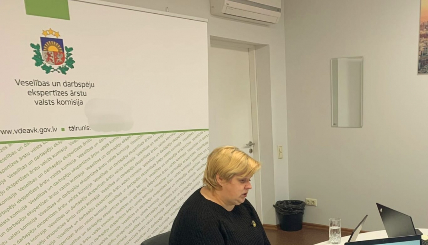 galds, pie galda sēž sieviete, uz galda dators, ūdens pudele, piezīmju grāmata